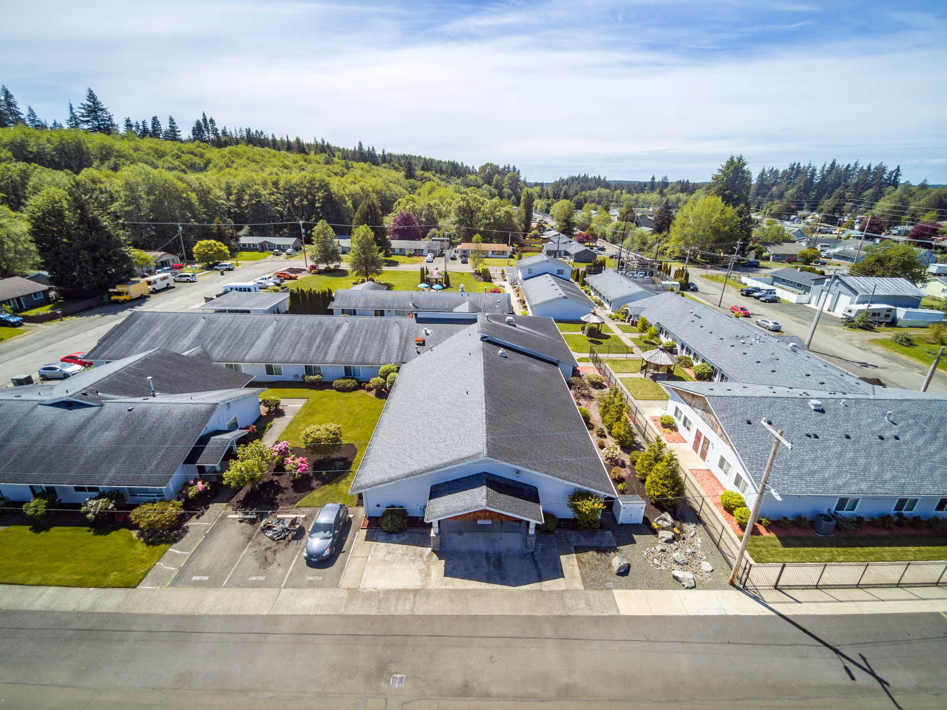 Aerial view of Beehive Retirement Community showing multiple single-story buildings with gray roofs arranged around green lawns and pathways, surrounded by trees and a residential neighborhood under a partly cloudy sky.