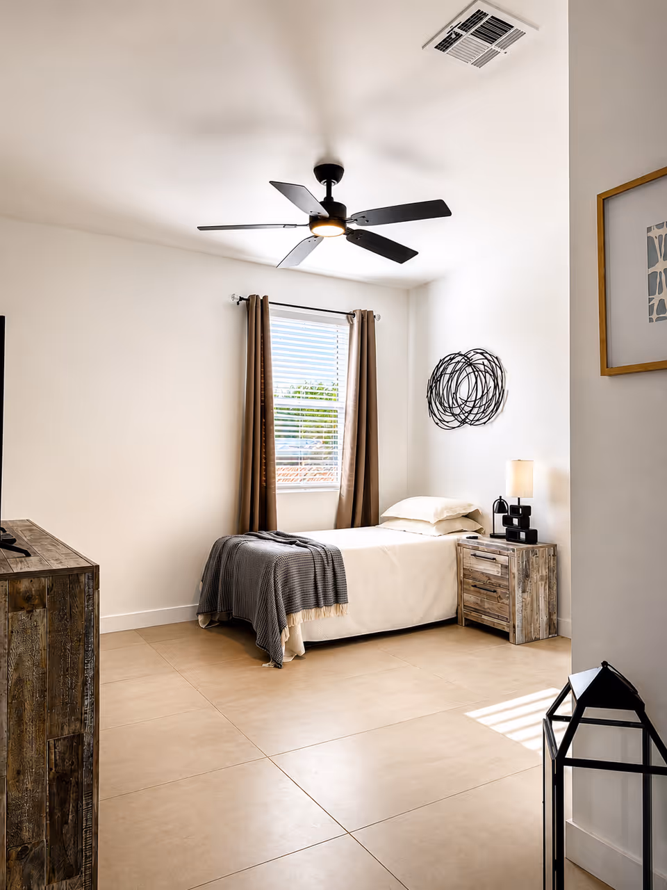 Bedroom in a senior living facility with bed, nightstand, window, and ceiling fan.