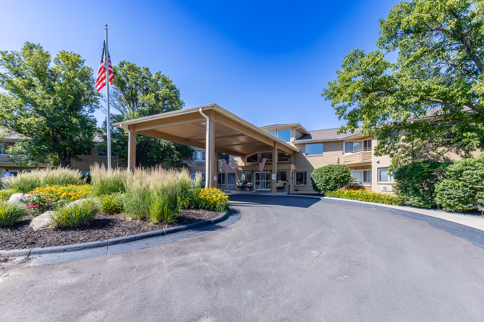 Exterior view of Asher Point Independent Living of Lincoln building entrance with a covered driveway, landscaped garden with flowers and tall grasses, an American flag on a flagpole, and trees under a clear blue sky.