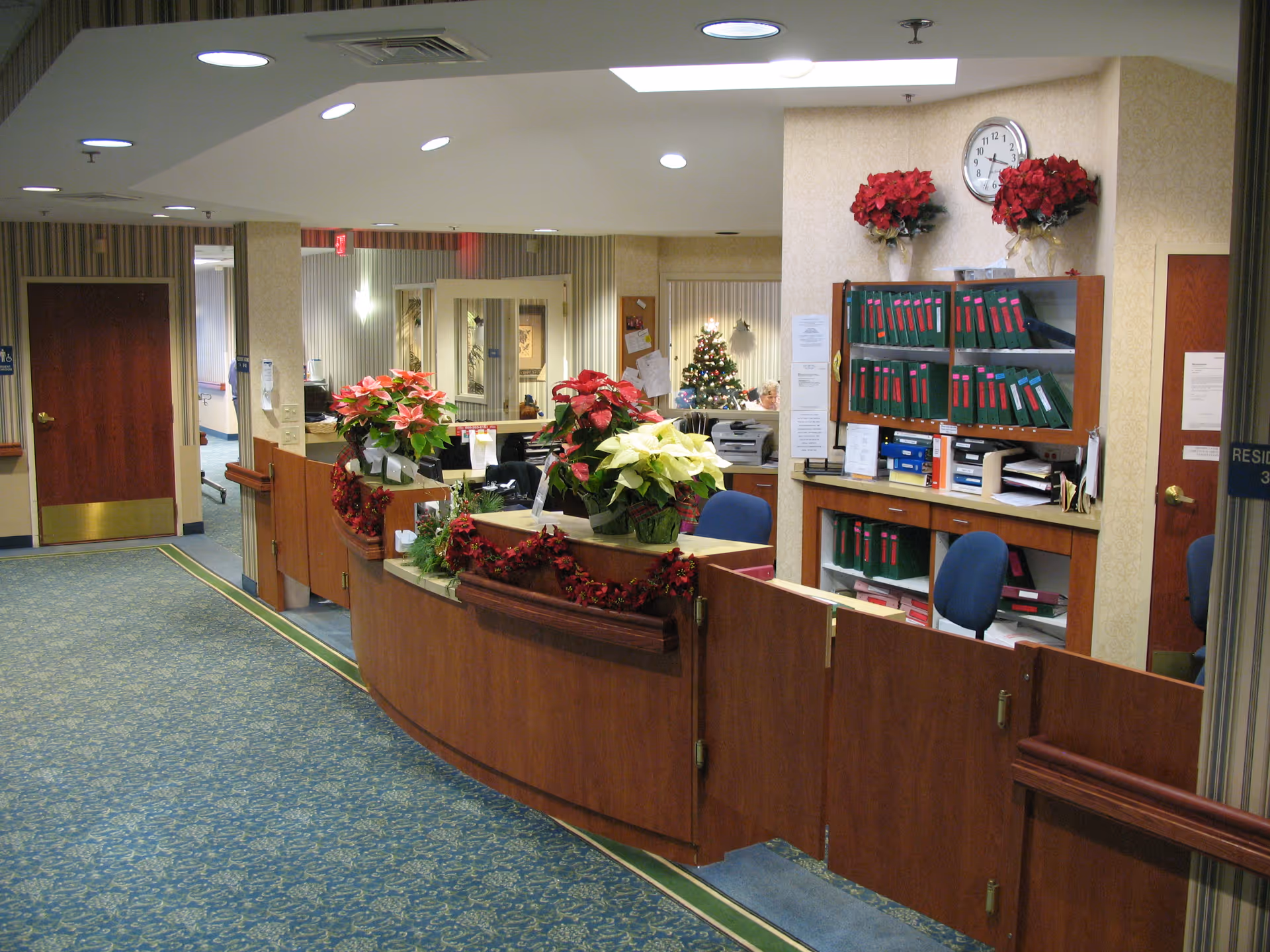 Reception area in a senior living facility with a curved wooden desk decorated with poinsettia plants and garlands. Behind the desk are shelves filled with green binders and office supplies. A clock and two poinsettia plants are mounted on the wall above the shelves. The carpet is blue with a floral pattern, and there are doors and hallways visible in the background.