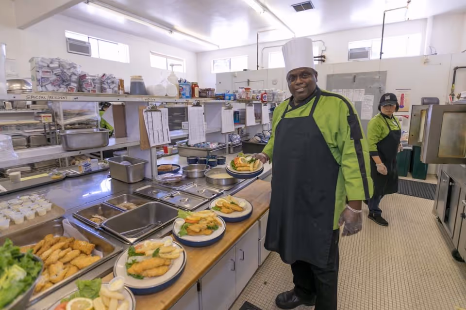 Two kitchen staff members in green uniforms and black aprons preparing and plating meals in a commercial kitchen. One person is wearing a chef's hat and holding a plate of food, while the other is standing near kitchen equipment in the background. The kitchen has stainless steel counters, food trays, and various kitchen supplies.
