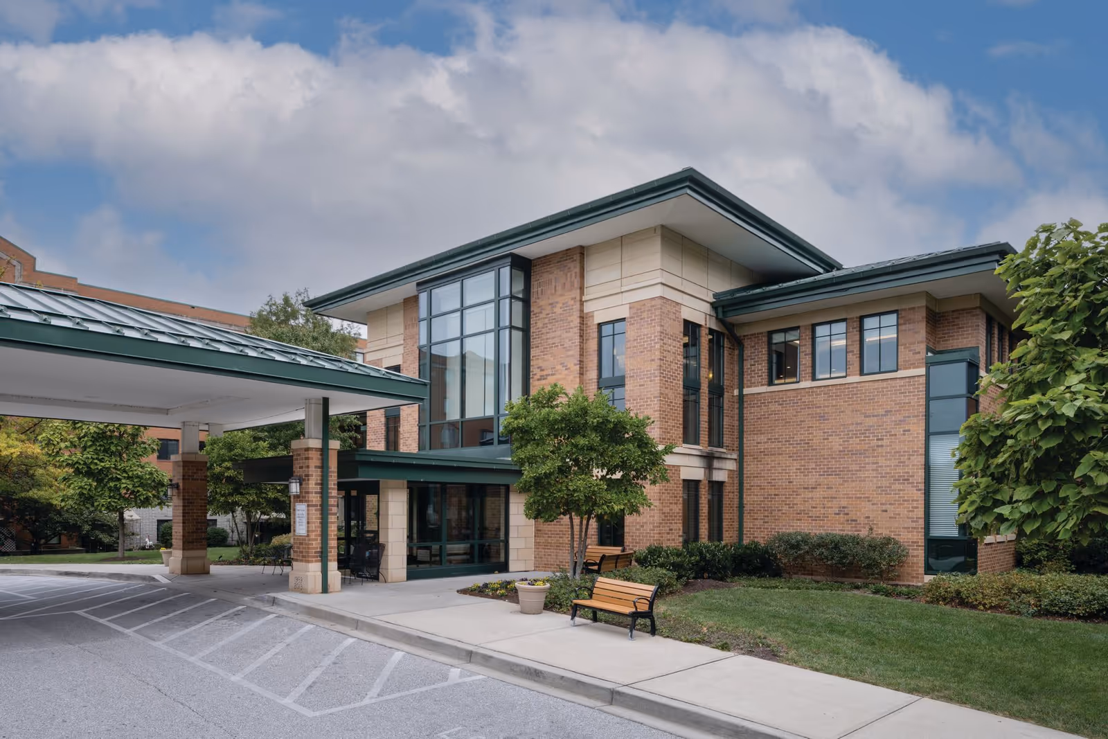 Brick senior living facility exterior with glass entrance, canopy, trees, and benches.
