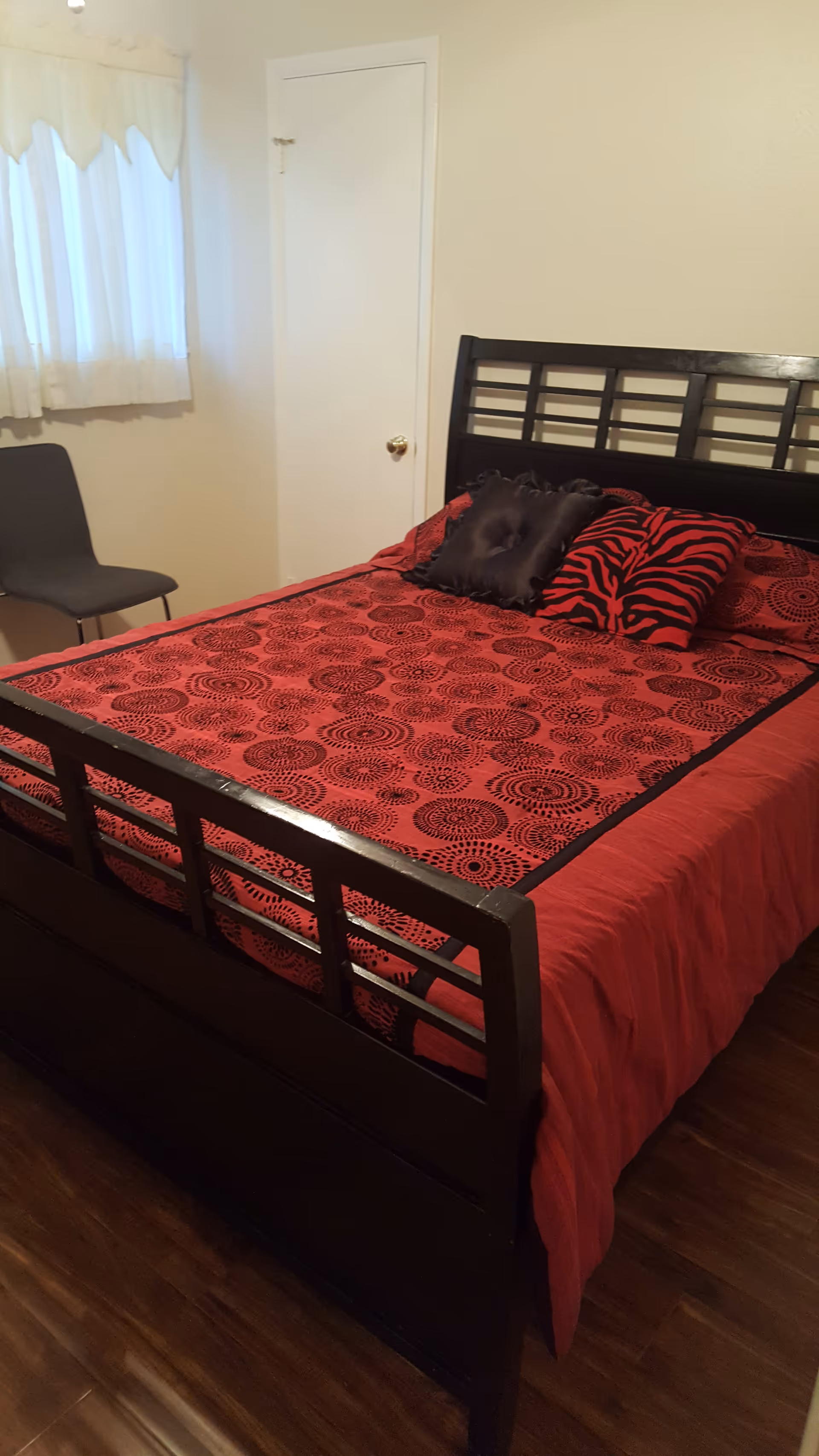 A neatly made bedroom with a dark wood bed, red patterned bedding, and a window chair.