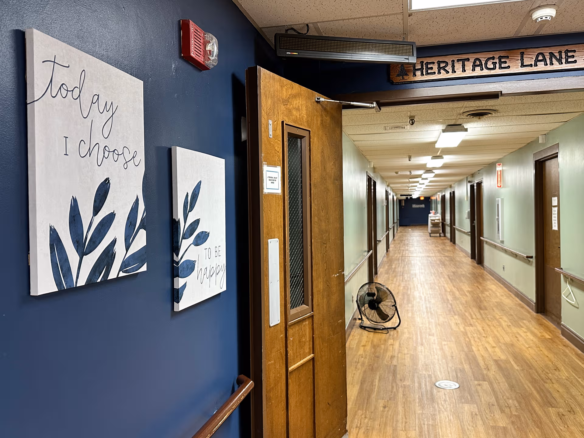 A long corridor with blue and green walls, doorway, wall art, and a floor fan.