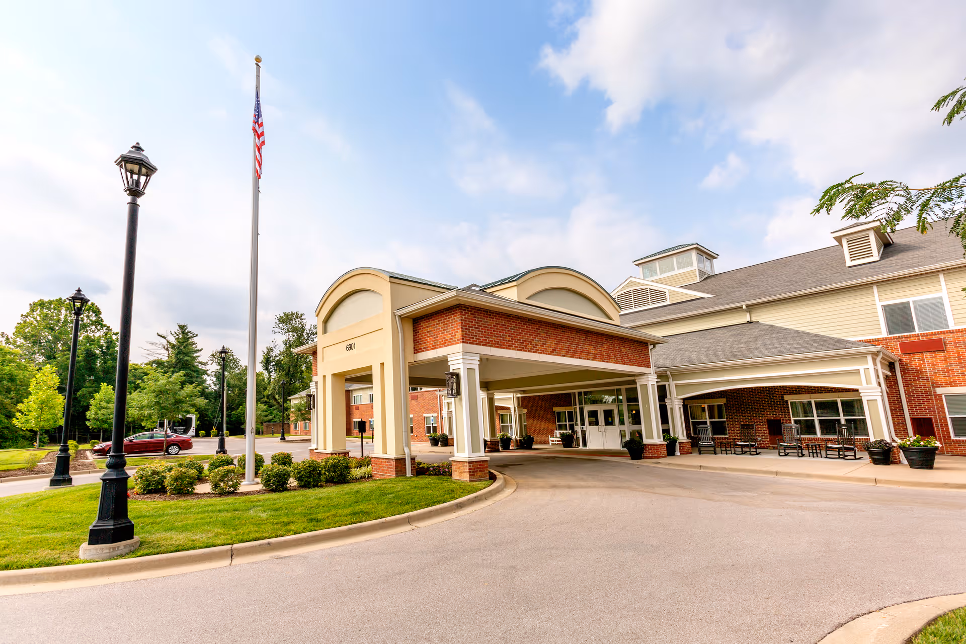 Covered entrance of a brick senior living building with a flagpole, driveway, and landscaping.