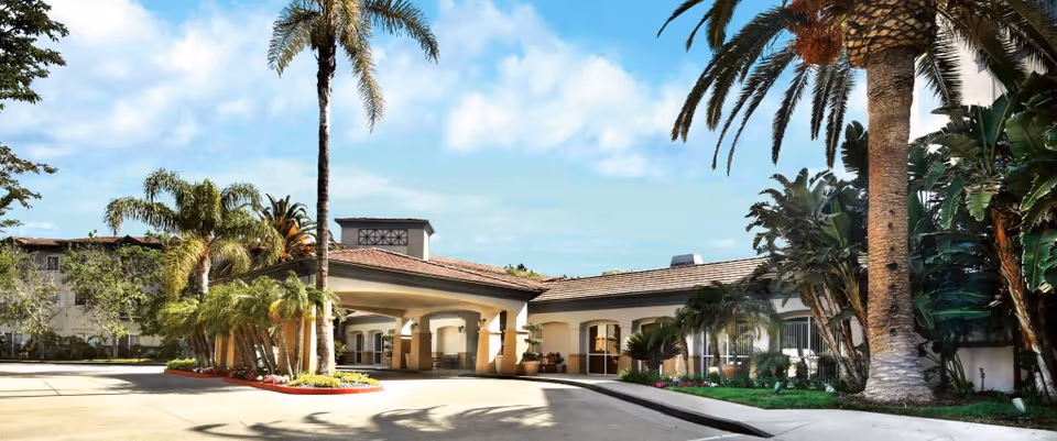 Exterior view of The Village at Sherman Oaks senior living facility featuring a covered entrance with arches, surrounded by palm trees and lush landscaping under a partly cloudy sky.