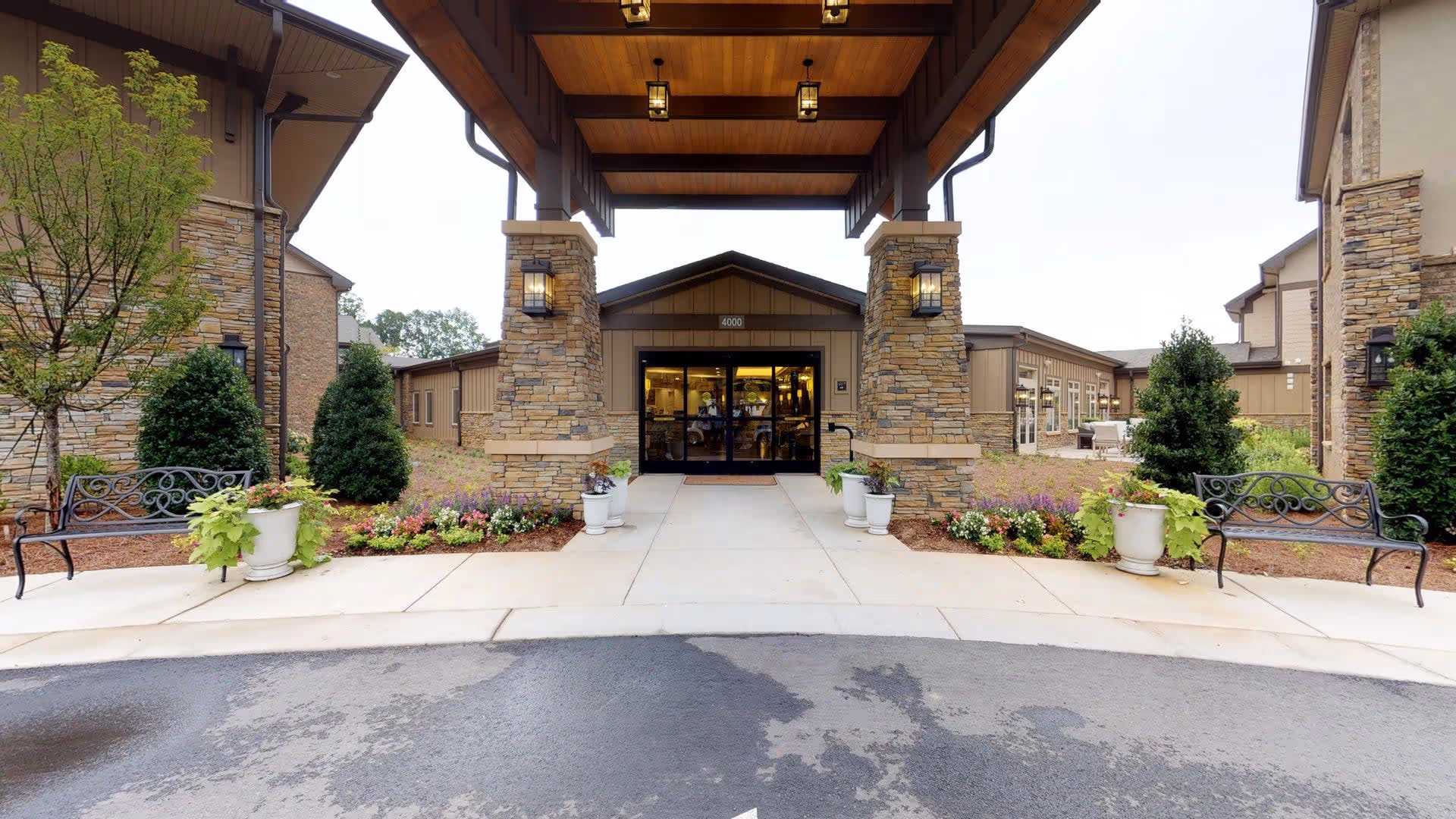 Entrance of Celebration Village Forsyth featuring a covered driveway with stone pillars, benches on either side, potted plants, and landscaped flower beds leading to glass double doors.