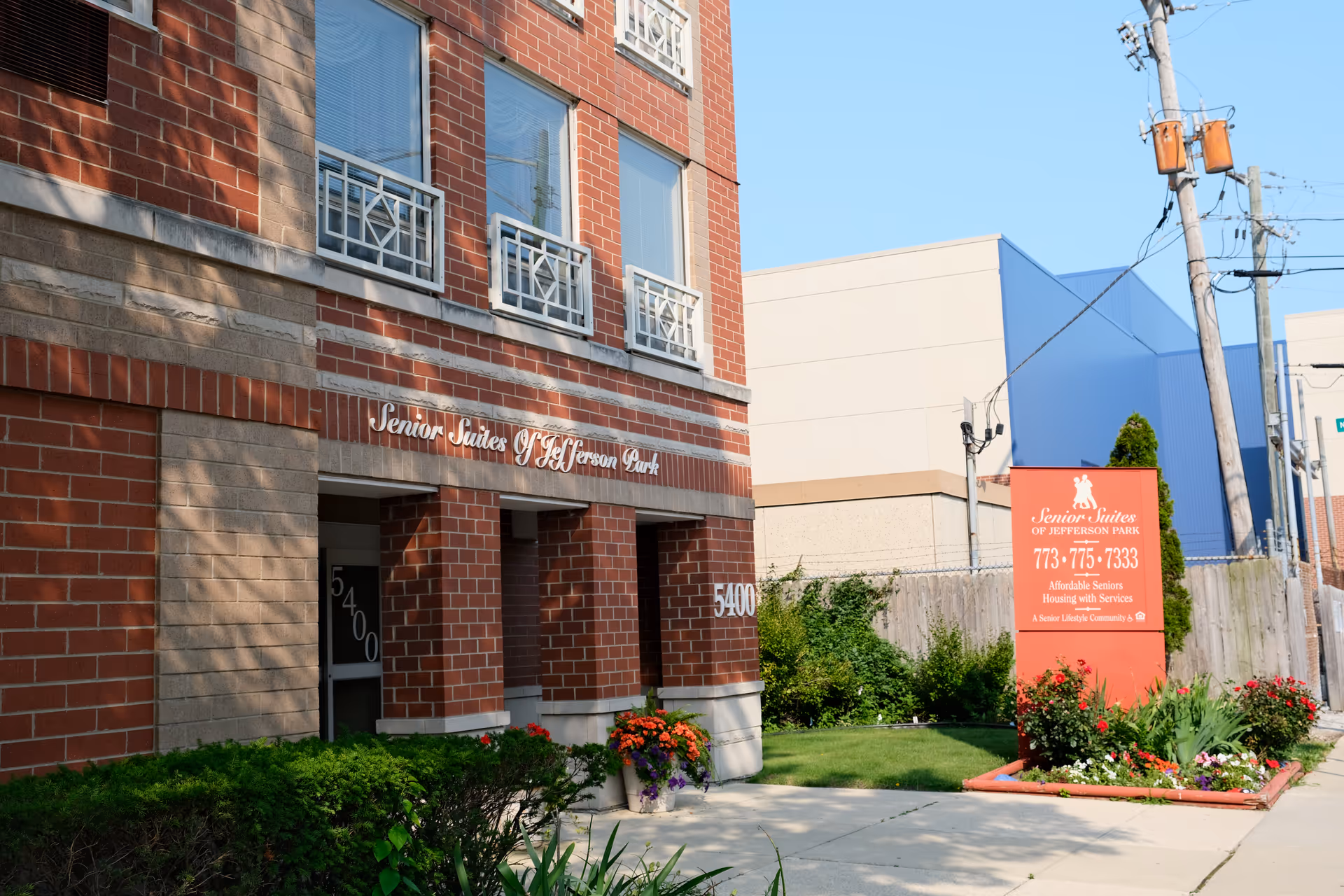 Exterior view of the Senior Suites of Jefferson Park building showing a brick facade with windows and a sign displaying the facility's name and contact information. There are plants and flowers near the entrance and a sidewalk leading to the building.