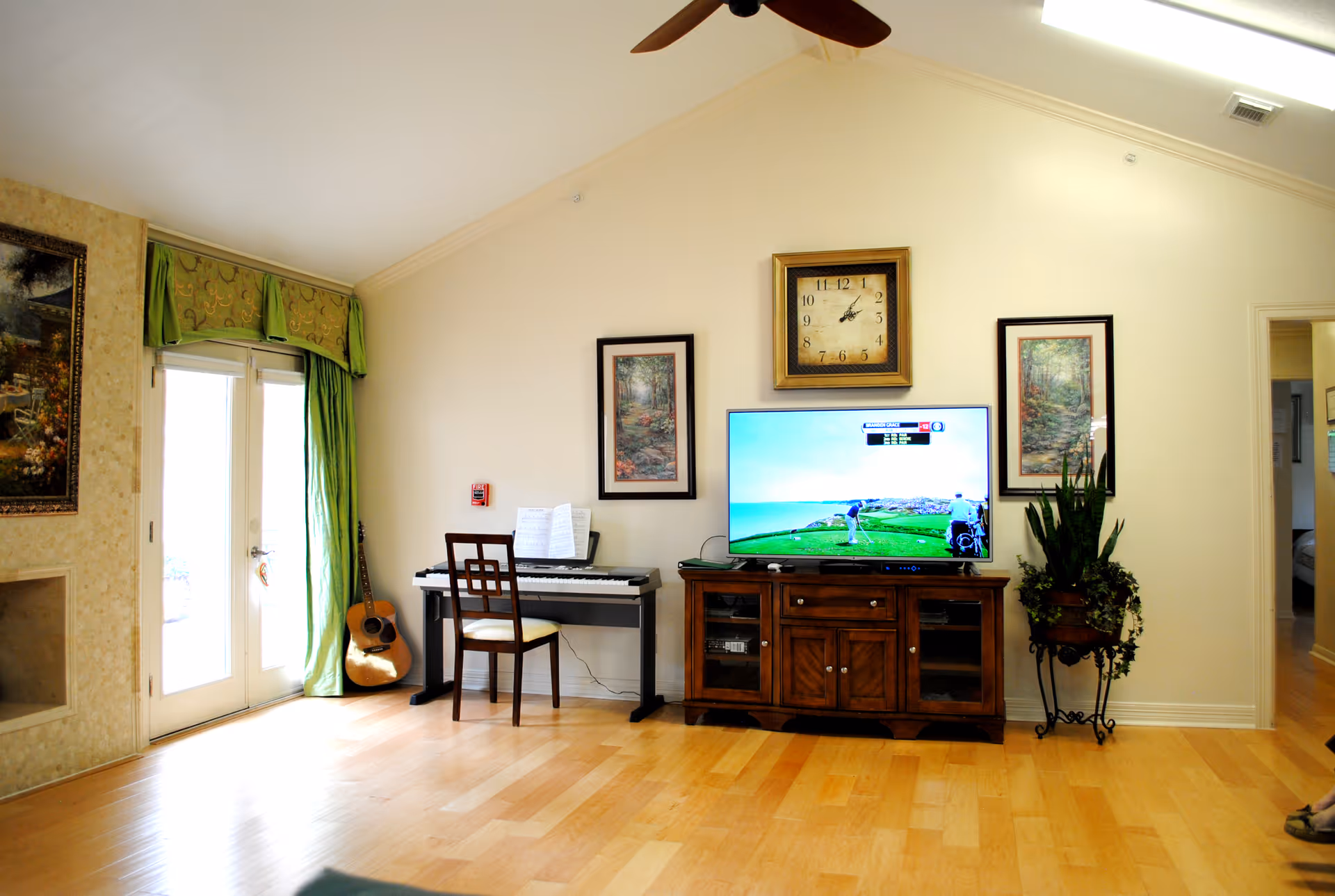 Bright living room with piano, guitar, TV stand, plants, and French doors in a senior facility.