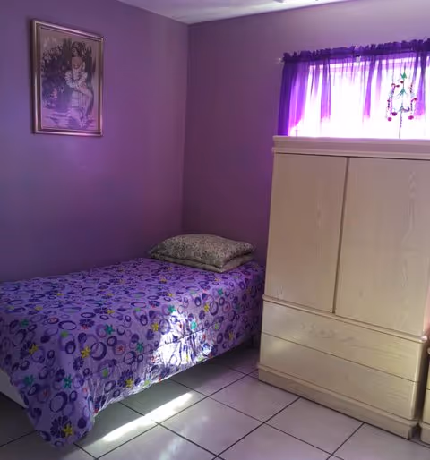 A small purple-walled bedroom with a single bed, dresser, wardrobe, and window.
