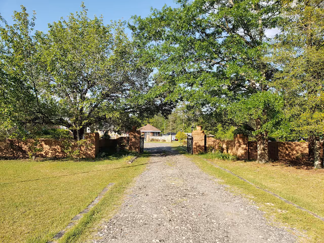 Gravel driveway flanked by brick walls and leafy trees leading to a distant facility entrance.
