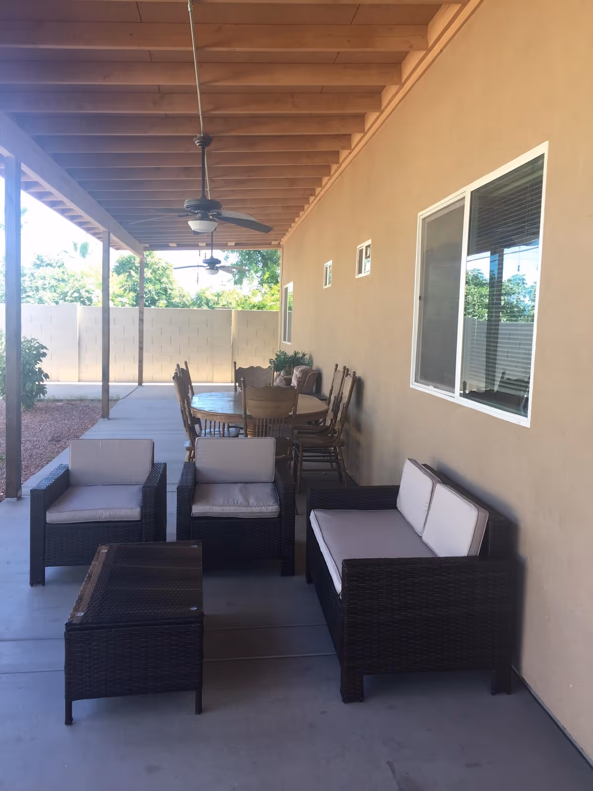Covered backyard patio with wicker seating, a coffee table, and a dining table under a wooden-beamed ceiling with ceiling fans.