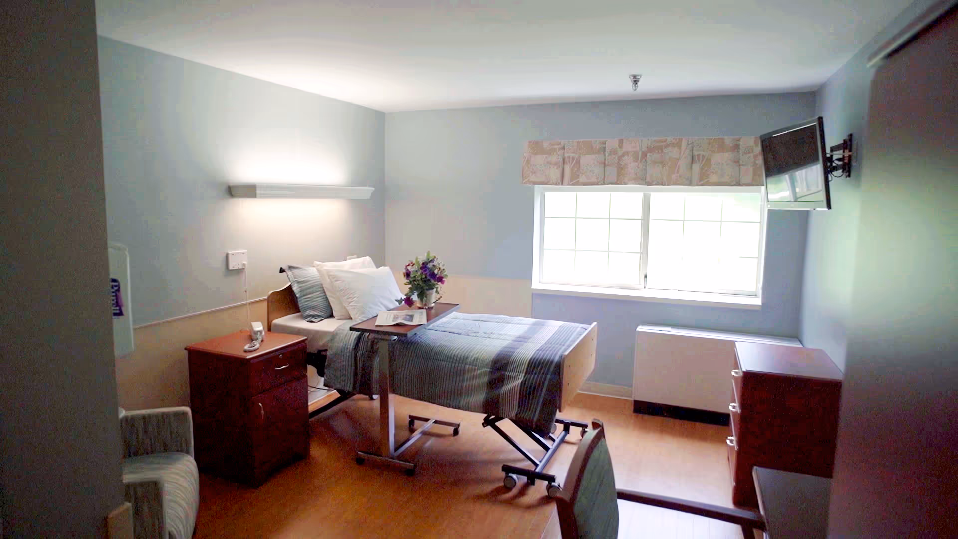 A bright nursing facility patient room with a single adjustable bed, bedside table, tray with flowers, window, dresser and wall-mounted TV.