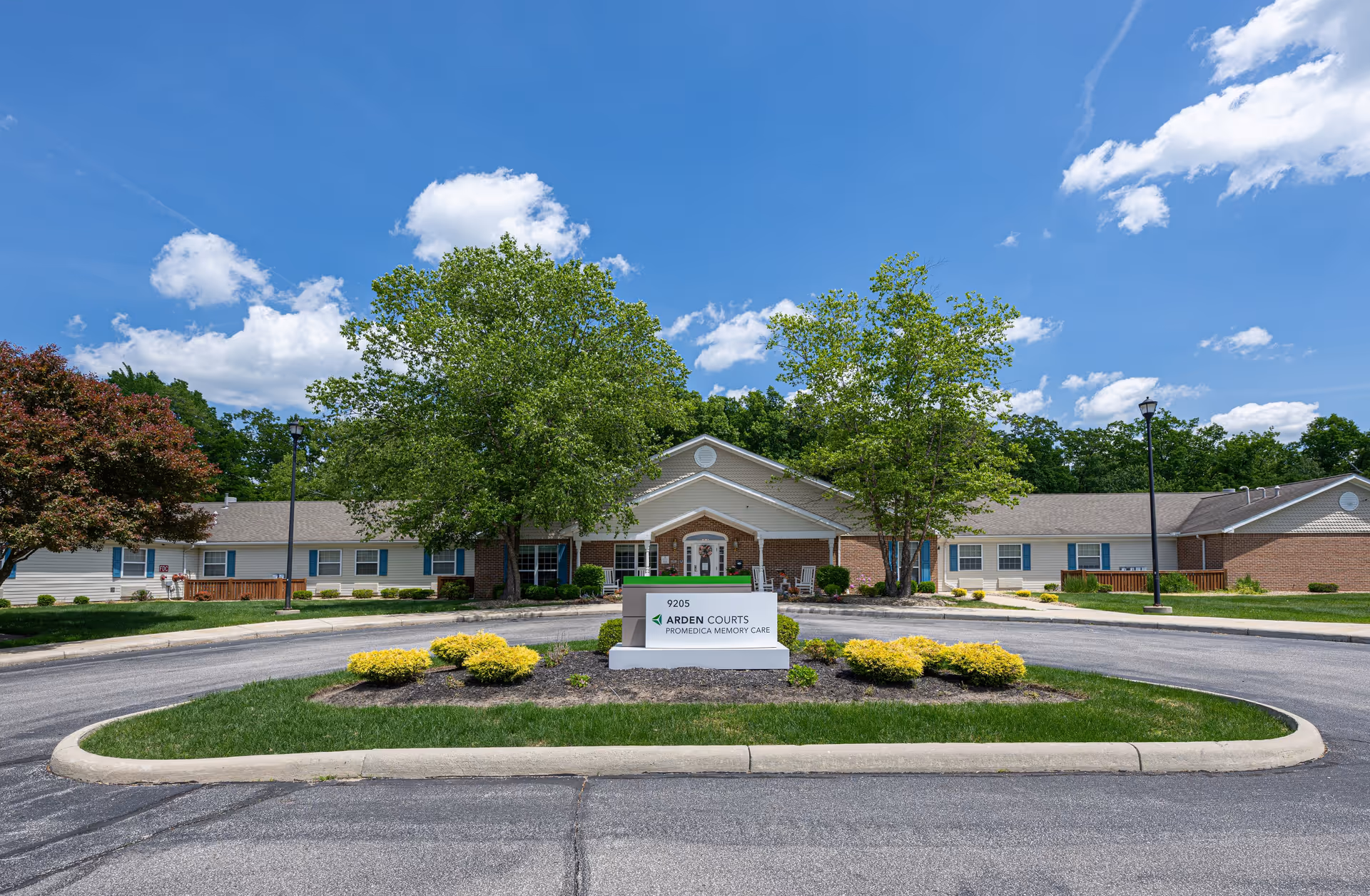 Front exterior view of Arden Courts - ProMedica Memory Care Community in Parma, showing a single-story building with a circular driveway, green trees, and a clear blue sky with some clouds.