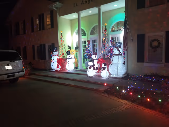 Front entrance of St. Angelus decorated at night with illuminated snowmen, Christmas trees, and holiday lights.
