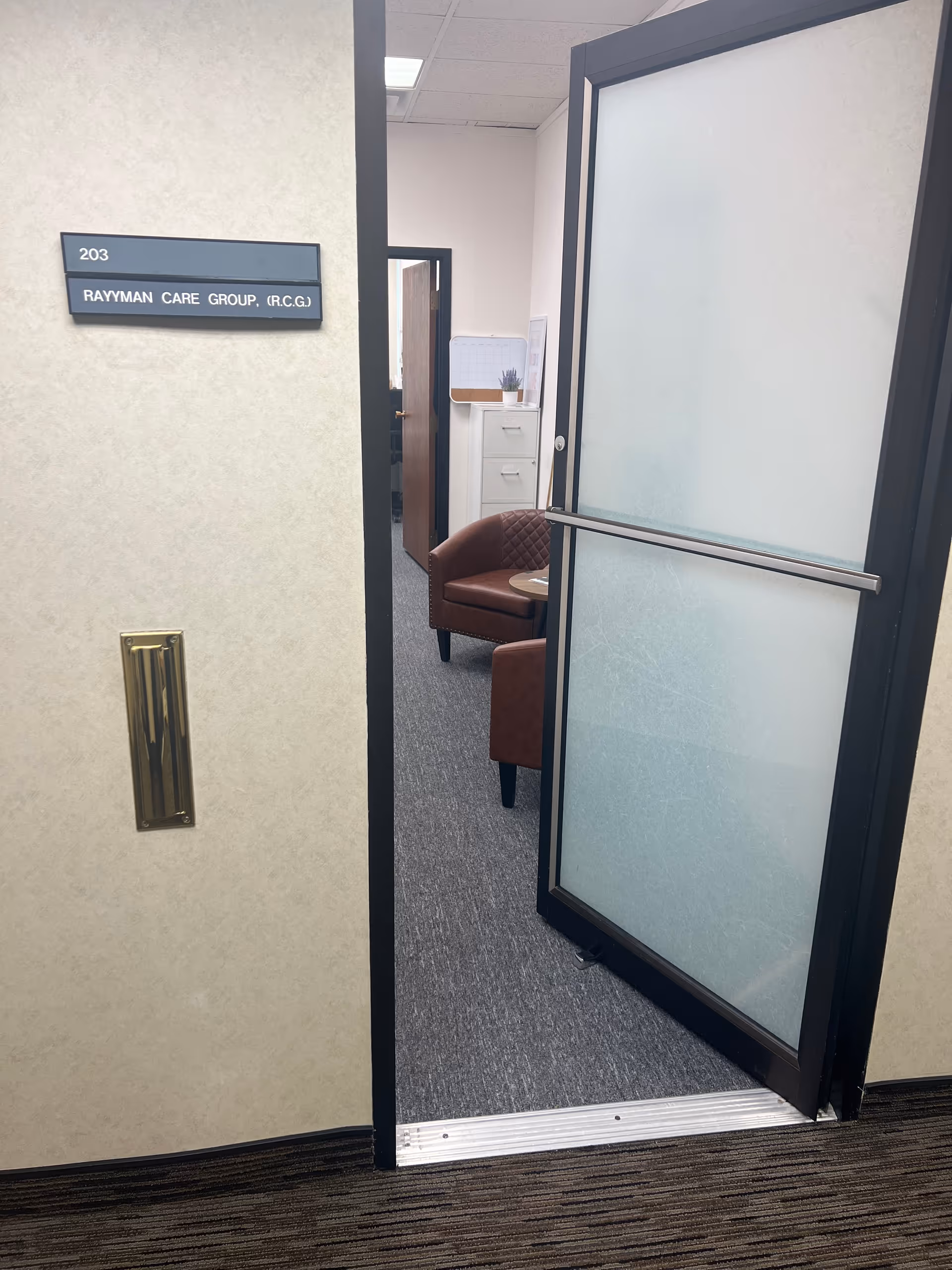 Open frosted glass door reveals waiting area with brown chairs and a sign.