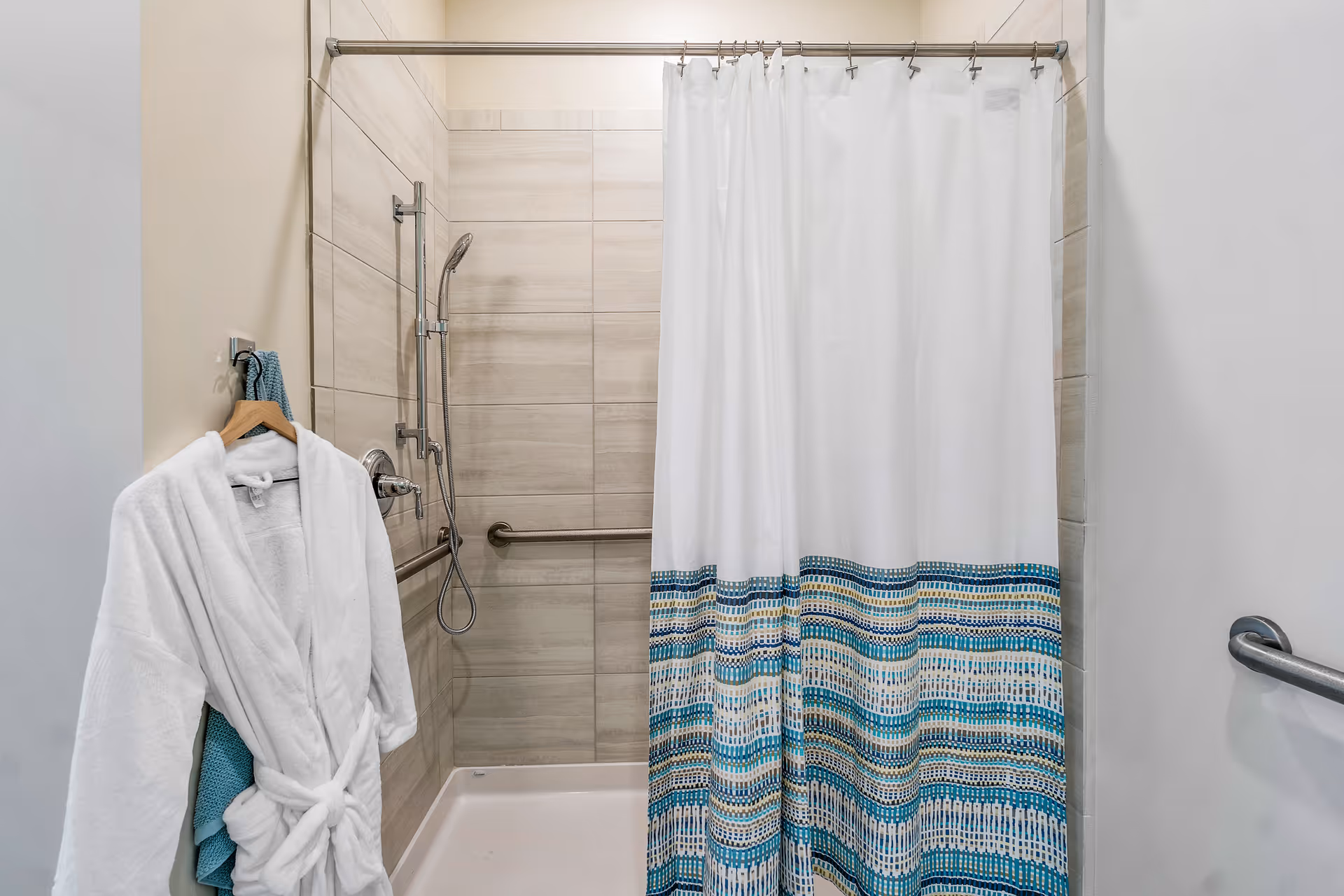 A bathroom shower area with beige tiled walls, a white shower curtain with blue and beige patterns at the bottom, a handheld showerhead, and a metal grab bar. A white bathrobe and a blue towel hang on a hook on the left wall.