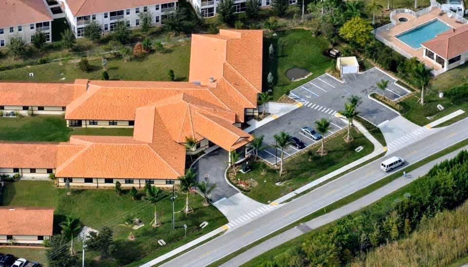 Aerial view of a senior living facility with orange-tiled roofs, parking lot, swimming pool, and a road with a van.