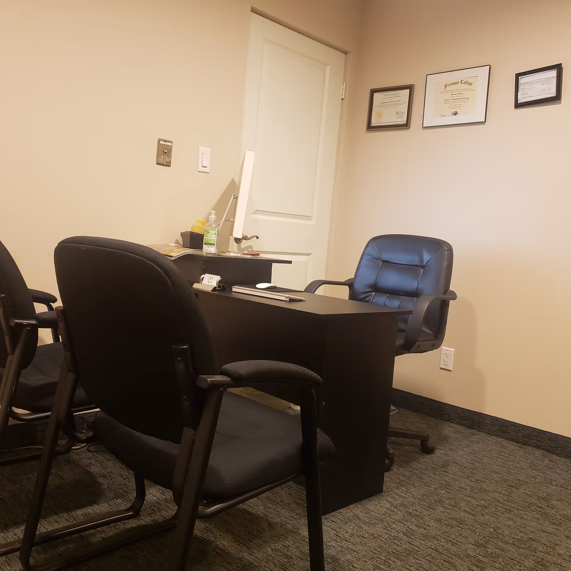 Office-like room with a desk, chairs, and framed certificates on beige walls.