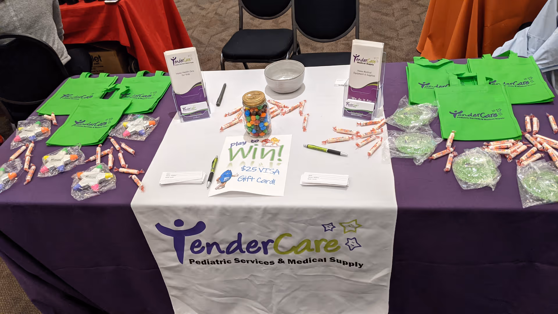 Indoors, a promotional table displays TenderCare branding, green tote bags, brochures, and wrapped candy.
