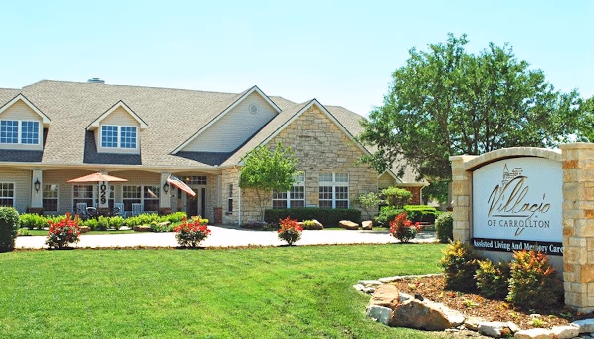 Exterior view of a single-story assisted living facility with a stone and siding facade, a well-maintained lawn, shrubs, and small trees. There is a sign in the foreground that reads 'Villacio of Carrollton Assisted Living And Memory Care.'