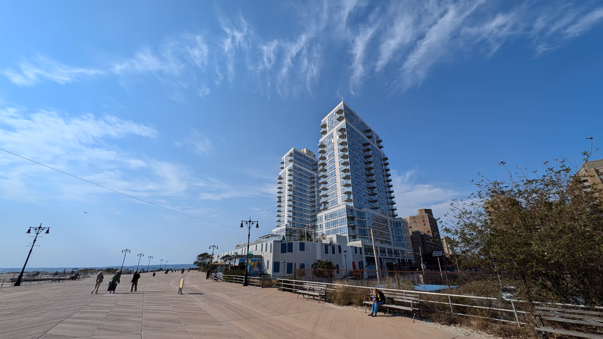 Tall modern seaside building with balconies overlooks a boardwalk with benches.