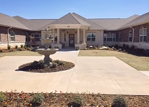 Front entrance of a single-story senior living building with a covered portico and a central tiered fountain in the driveway.