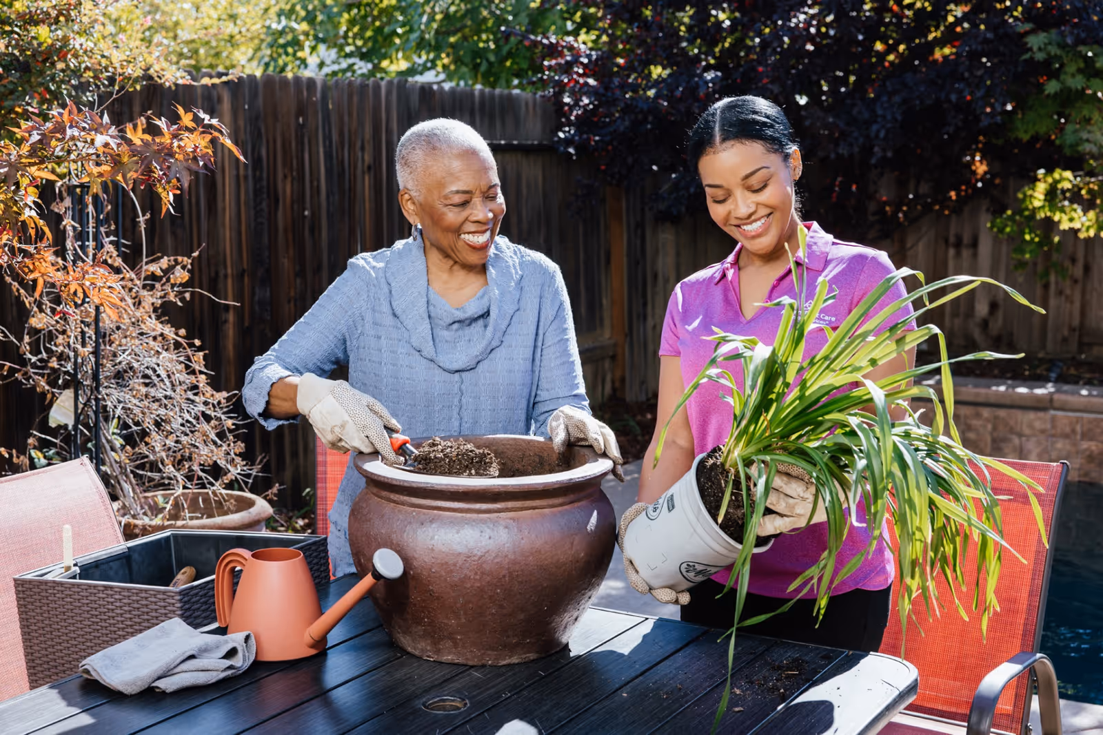 Two caregivers pot plants at an outdoor table, smiling in a sunny backyard.