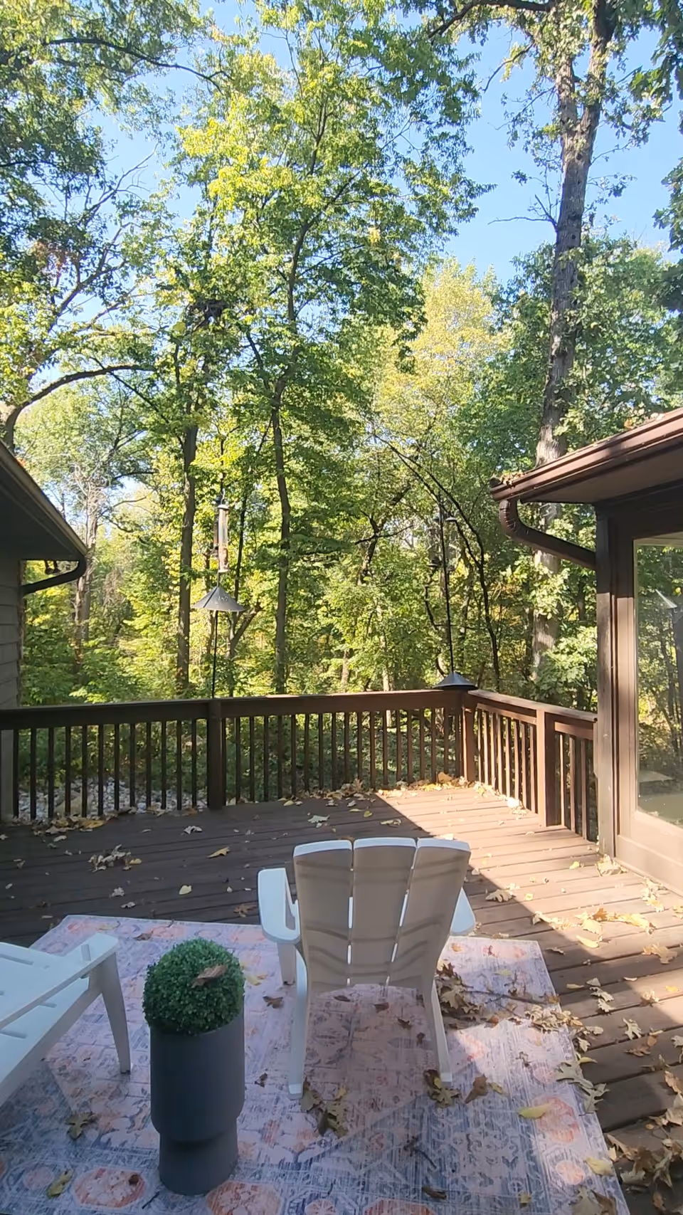 Outdoor wooden deck with plastic chairs, a table, potted plant, and surrounding trees.