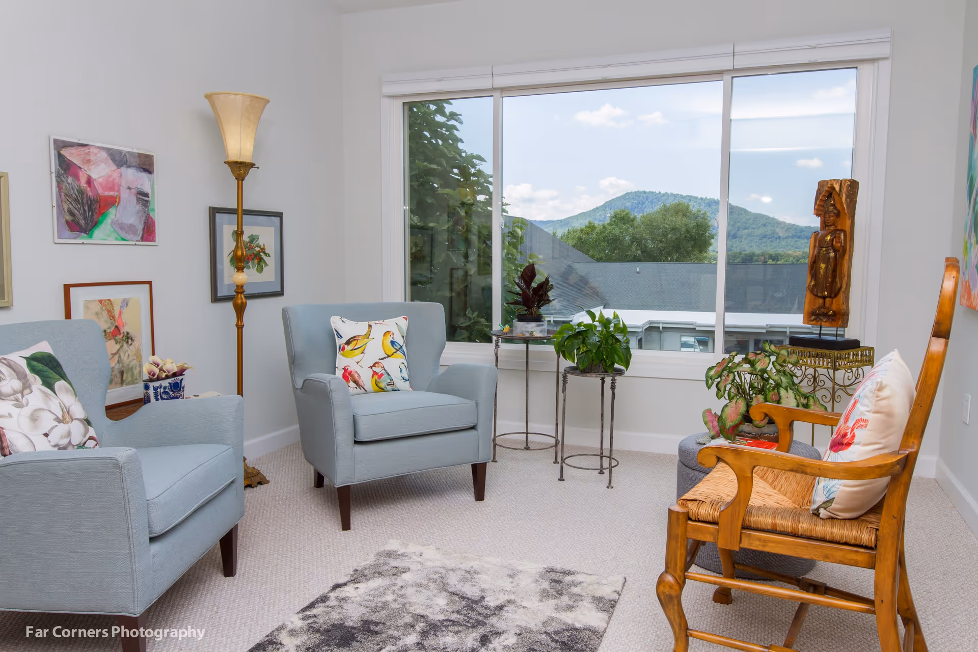 Bright seating area with two blue armchairs, a wooden chair, plants, artwork, and a large window showing a mountain view.