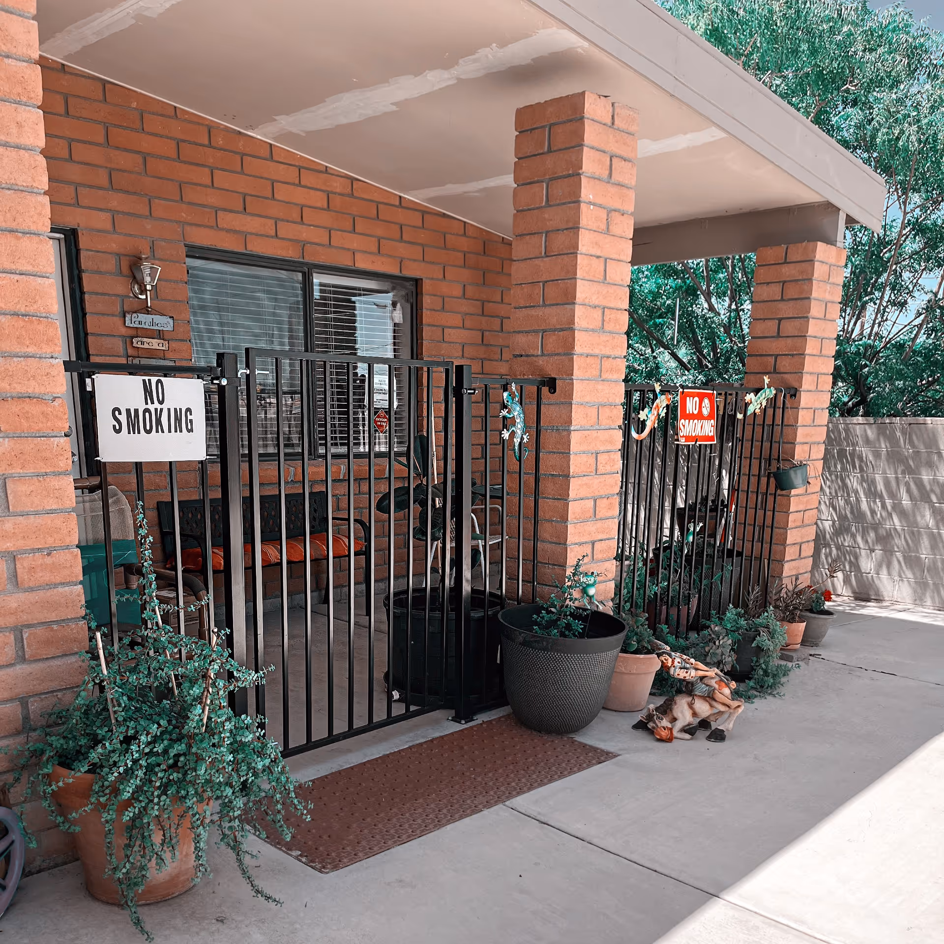 Outdoor patio area with brick pillars and a black metal gate featuring 'No Smoking' signs. Several potted plants and decorative items are placed around the gate. A bench with cushions is visible behind the gate, and trees are in the background.
