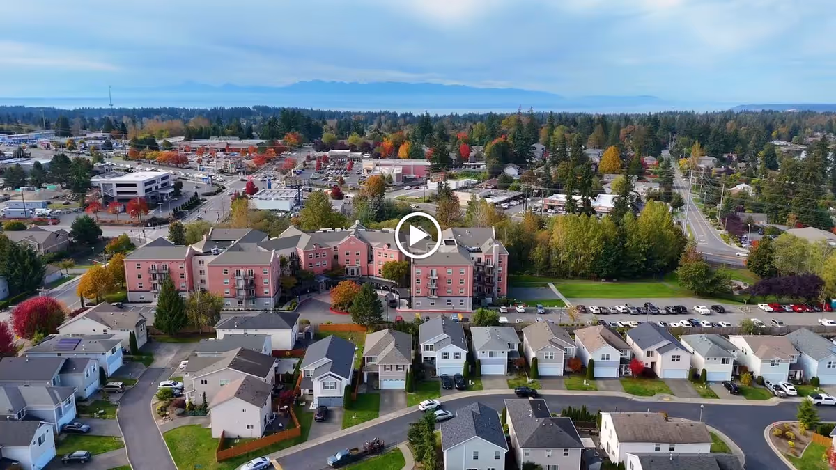 Aerial view of a pink multi-story retirement building surrounded by suburban houses, parking lots, and tree-lined streets with a distant horizon.