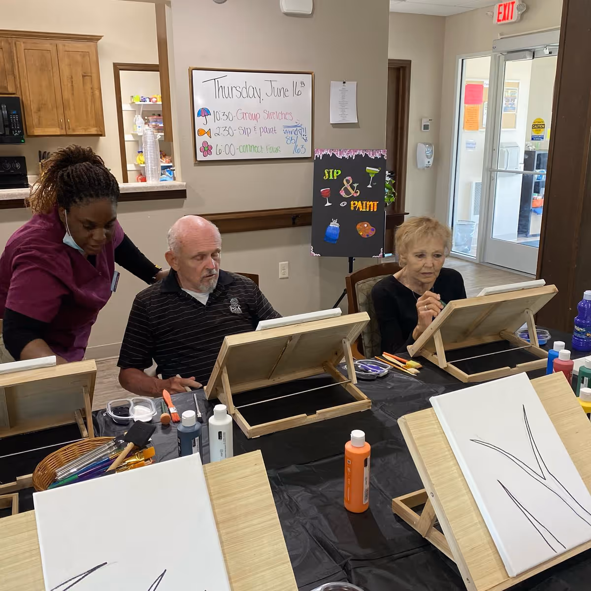 An elderly man and woman seated at a table with easels and painting supplies, participating in a sip and paint activity. A caregiver in a purple uniform is assisting the man. Behind them is a whiteboard with the day's schedule and a colorful sign that reads 'Sip & Paint'. The room appears to be a common area in an assisted living facility.