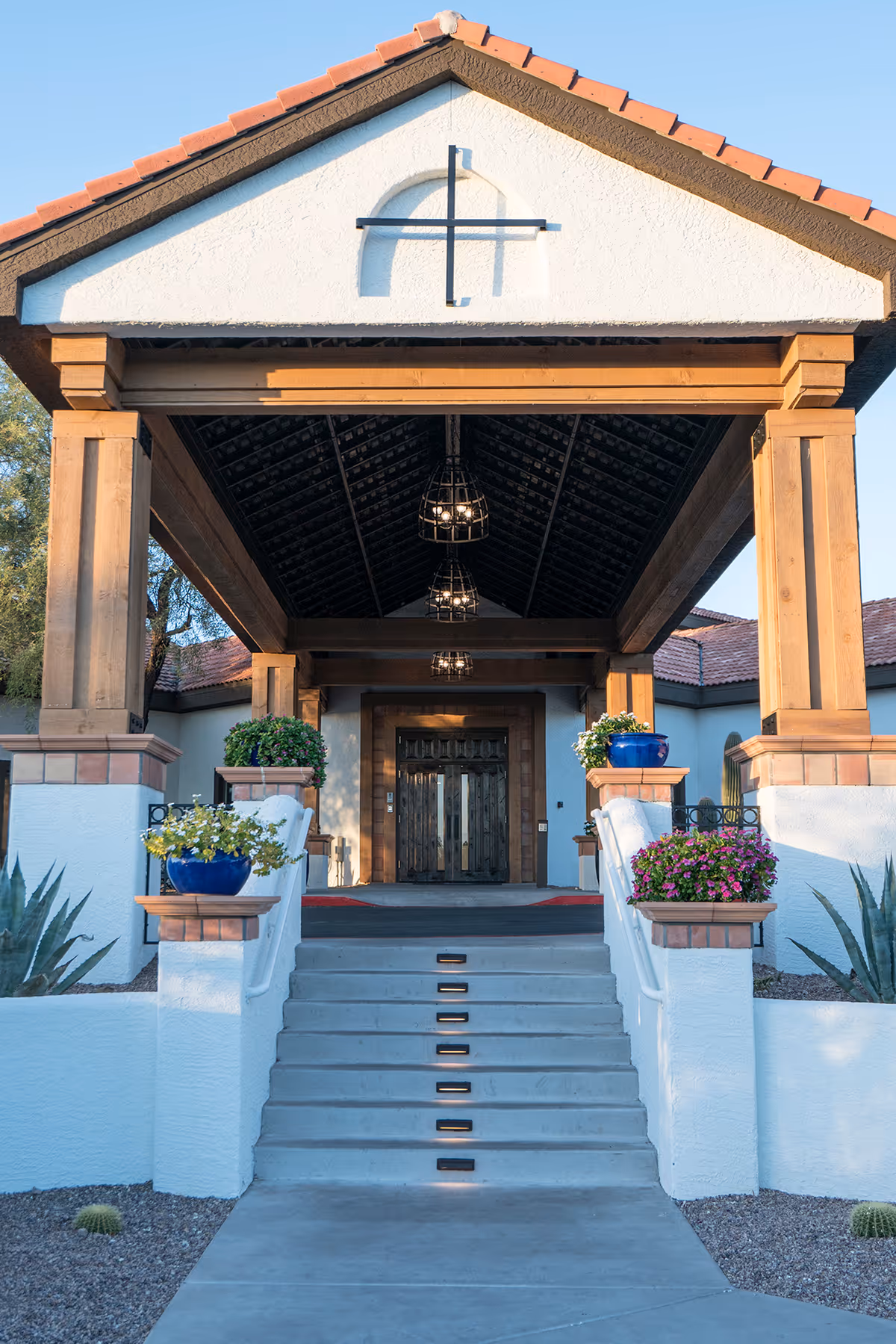 Gabled covered entrance with wooden columns and hanging lights, stairs leading up to double wooden doors flanked by potted flowers.