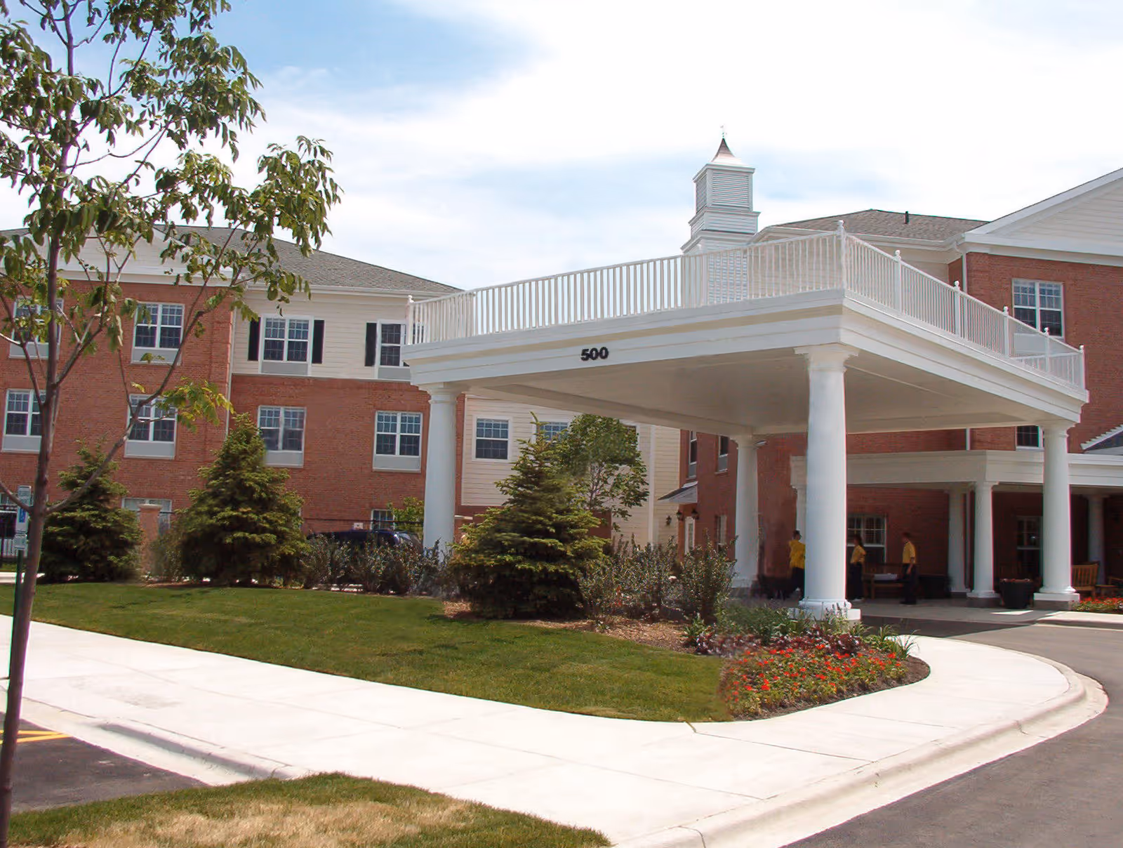 Exterior view of Belmont Village Senior Living Buffalo Grove showing a red brick building with white trim, a covered entrance supported by large white columns, landscaped greenery, and a clear sky.
