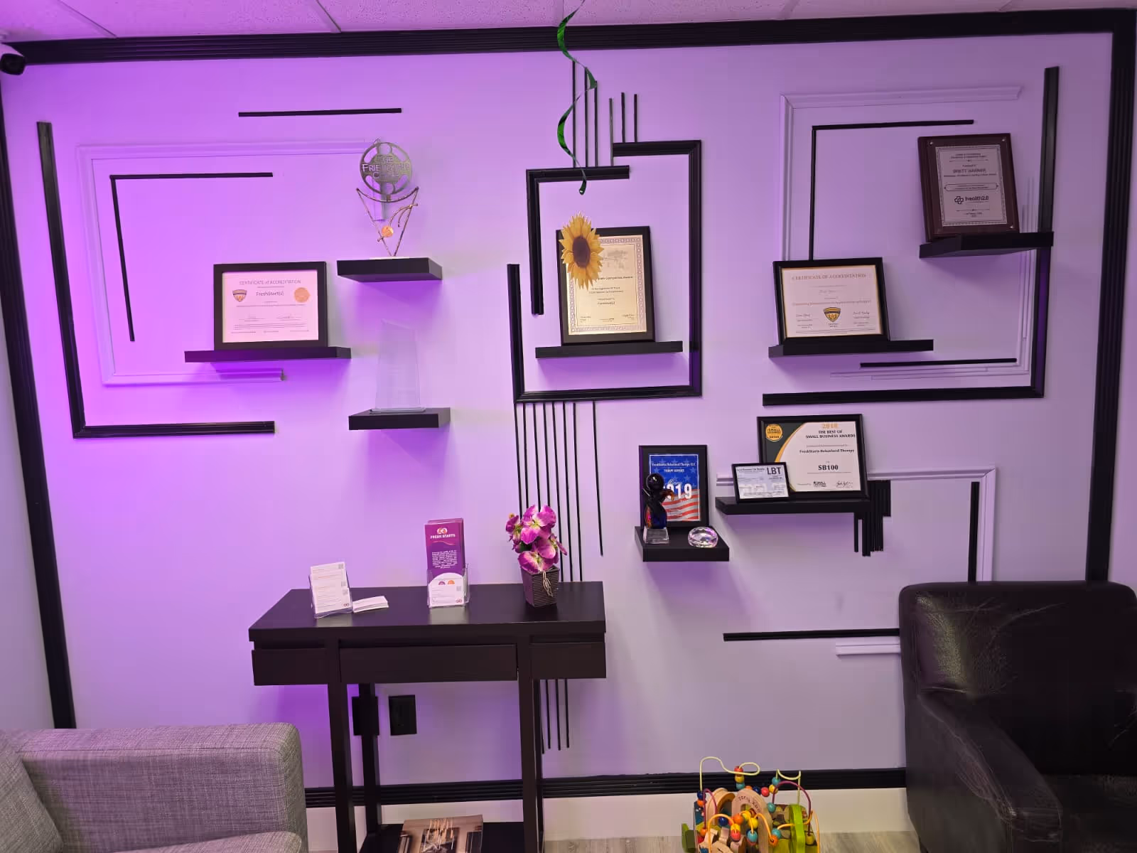 Purple-lit office wall with geometric black shelves, framed certificates, a desk, and a small toy.