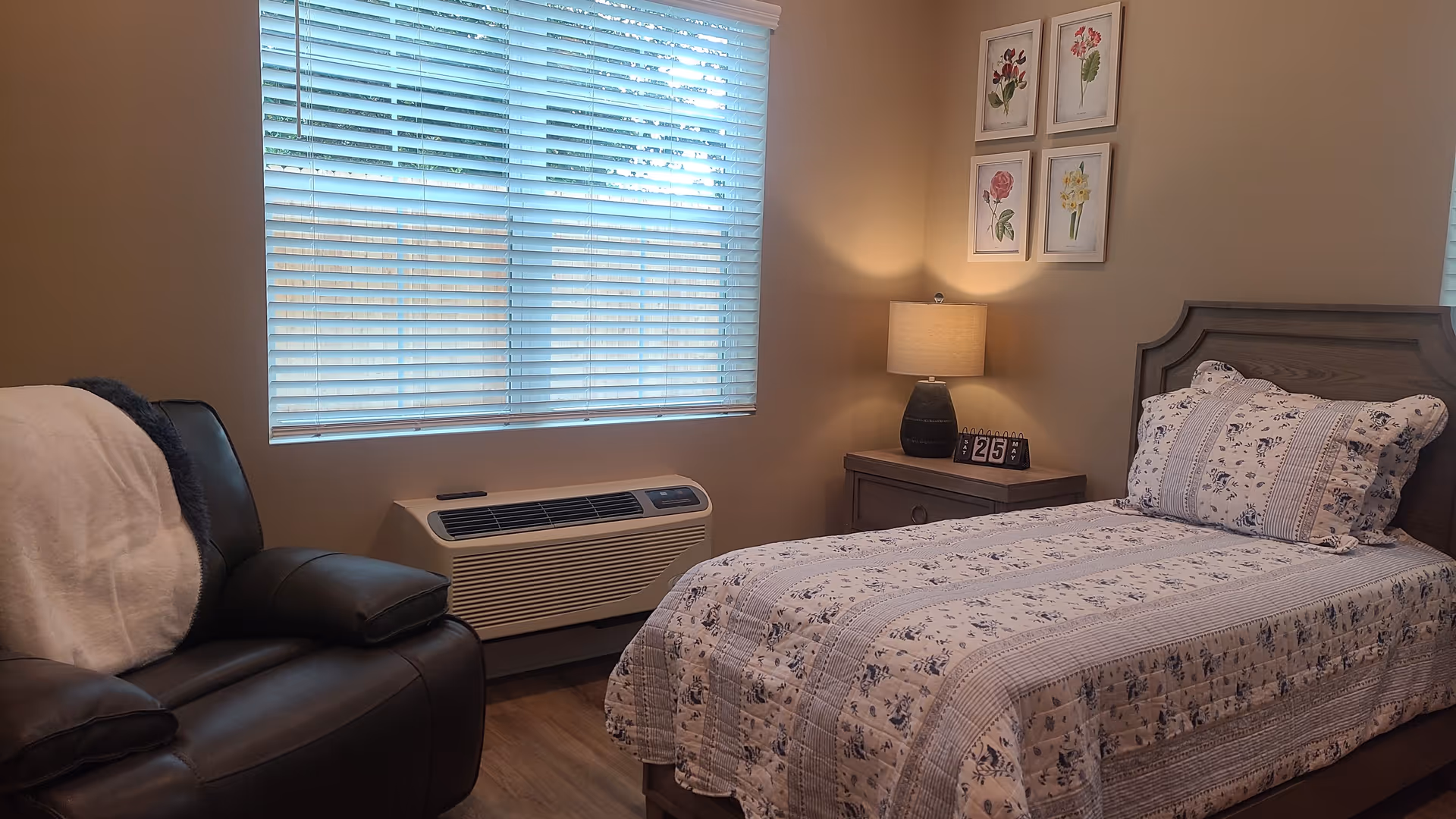 A single bedroom with bed, nightstand, lamp, recliner, and window blinds.