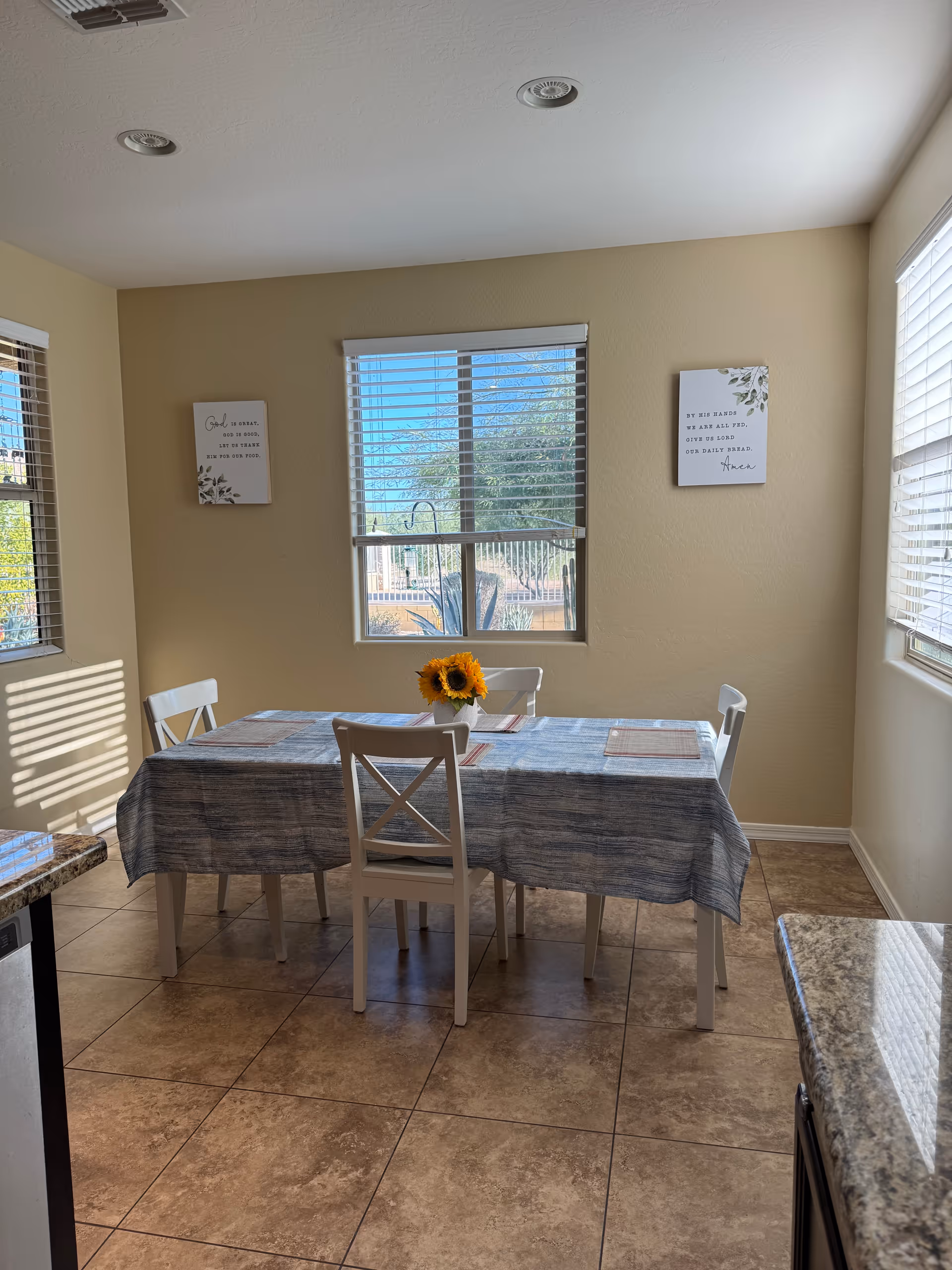 A bright dining area with a four-seat table, sunflower centerpiece, windows, and beige walls.