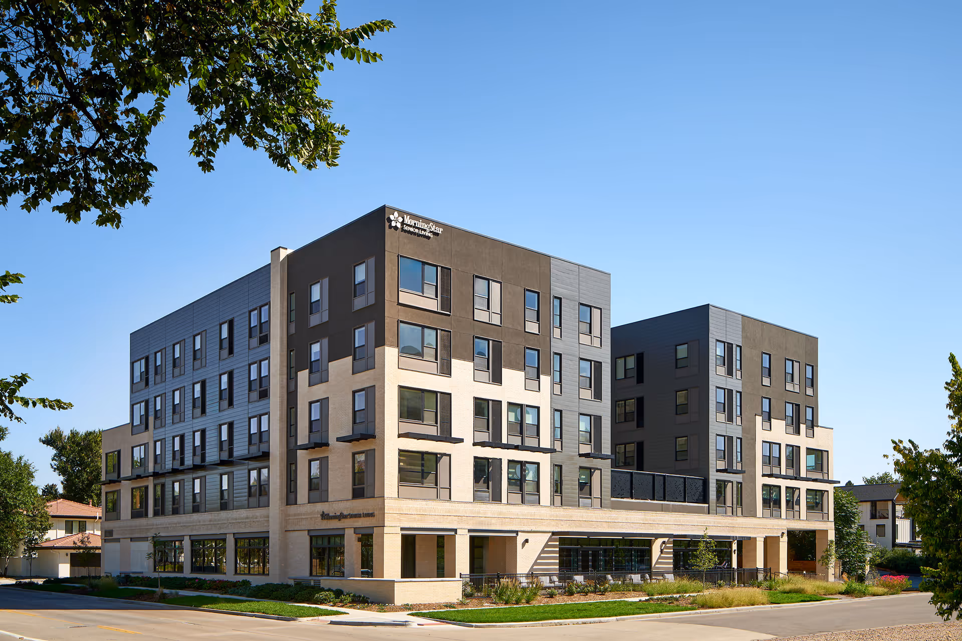 Modern multi-story assisted living building with large windows and landscaped frontage under a clear blue sky.