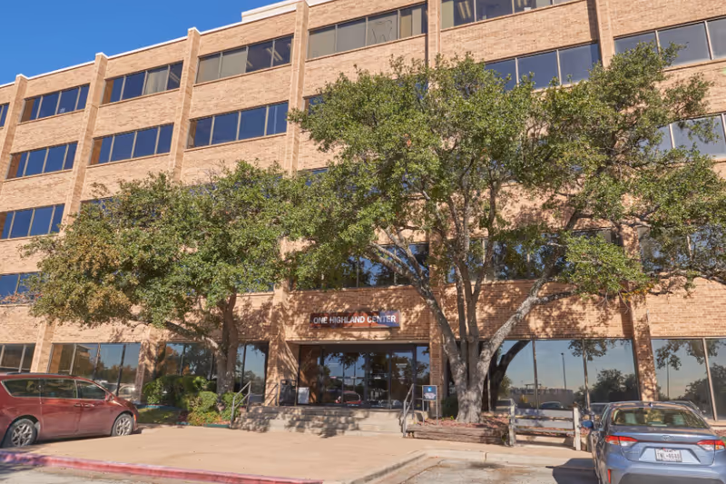 Front view of a brick building with large windows, trees, and a parking lot.