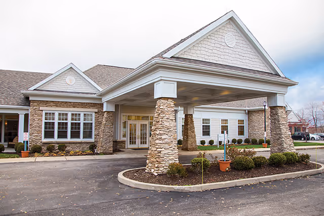 Front entrance of a senior living building with a covered porte-cochere supported by stone columns and landscaped planters.