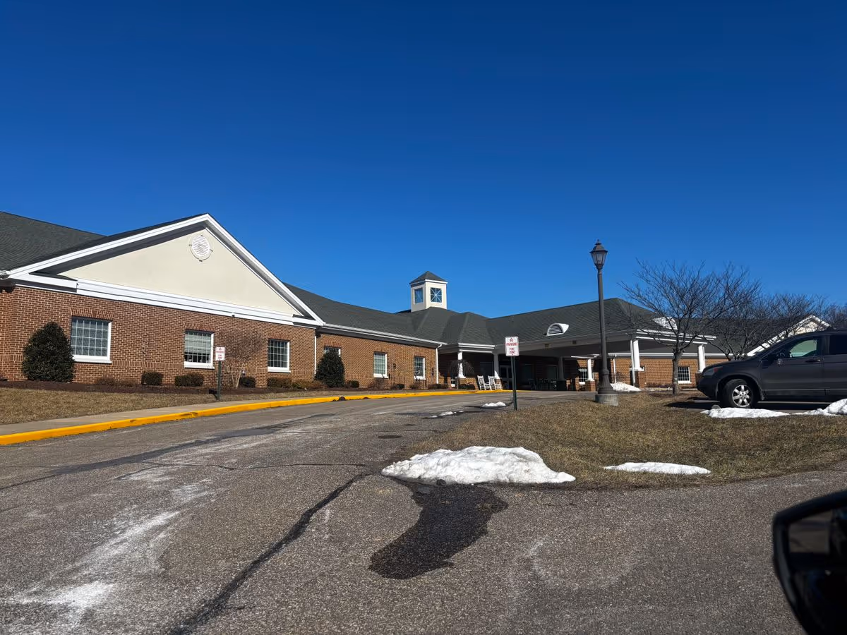 Front exterior of a single-story brick senior living building with a covered entrance, circular drive, lamp post, and small patches of snow.