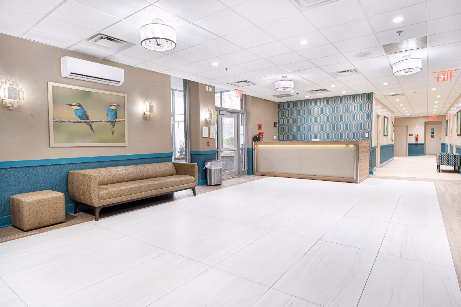 A spacious and well-lit reception area in a healthcare and rehabilitation center featuring a modern reception desk with a patterned blue wall behind it, a brown cushioned sofa and ottoman, a large framed picture of two colorful birds on a branch, ceiling lights, and a tiled floor. There are glass doors leading outside and a hallway extending to the right.