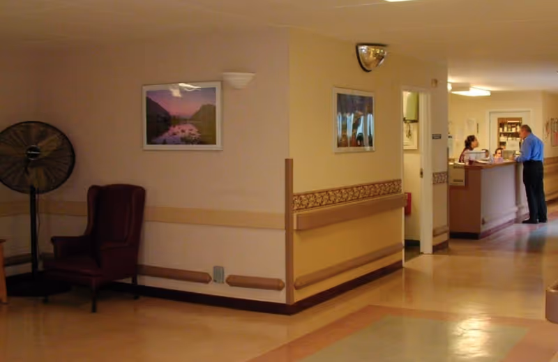 Interior hallway of a senior living facility with a reception desk at the far end, an armchair and framed art along the corridor.