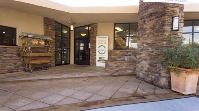 Covered stone-clad entrance to a senior living center with double glass doors, a decorative cart, potted plant, and a freestanding sign.