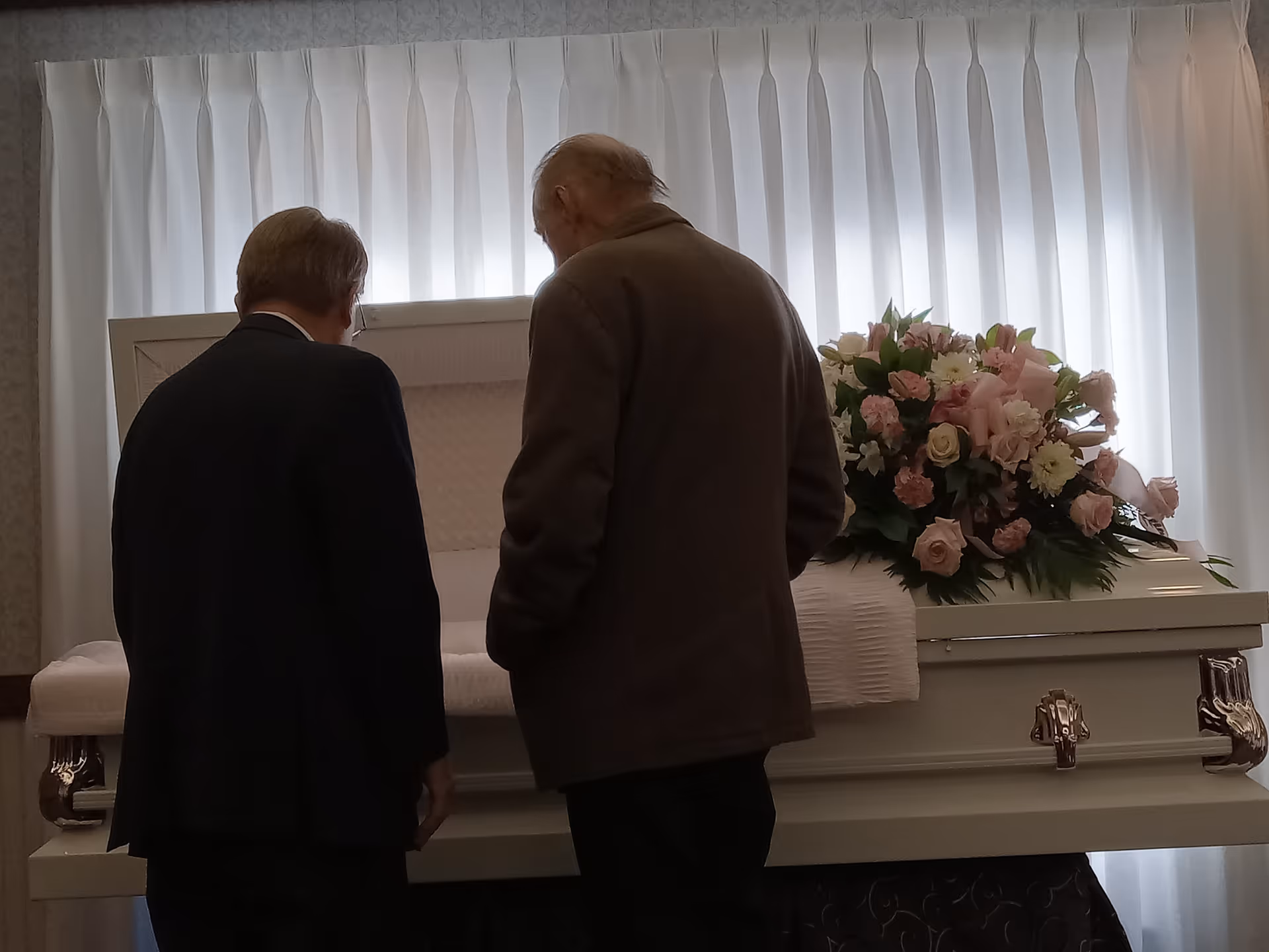 Two elderly men stand beside a white casket with pink flowers indoors.
