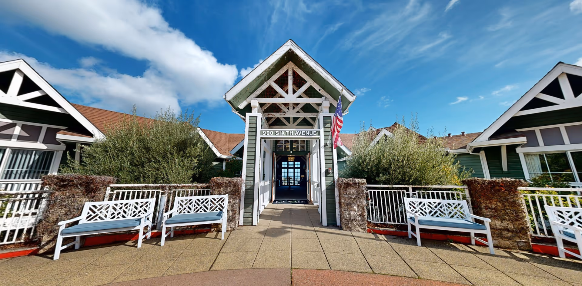 Entrance of a senior living facility with a peaked roof structure displaying the address 900 Sixth Avenue. The building is green with white trim and has an American flag hanging on the right side. There are white benches with blue cushions on either side of the entrance, surrounded by greenery and bushes under a partly cloudy blue sky.