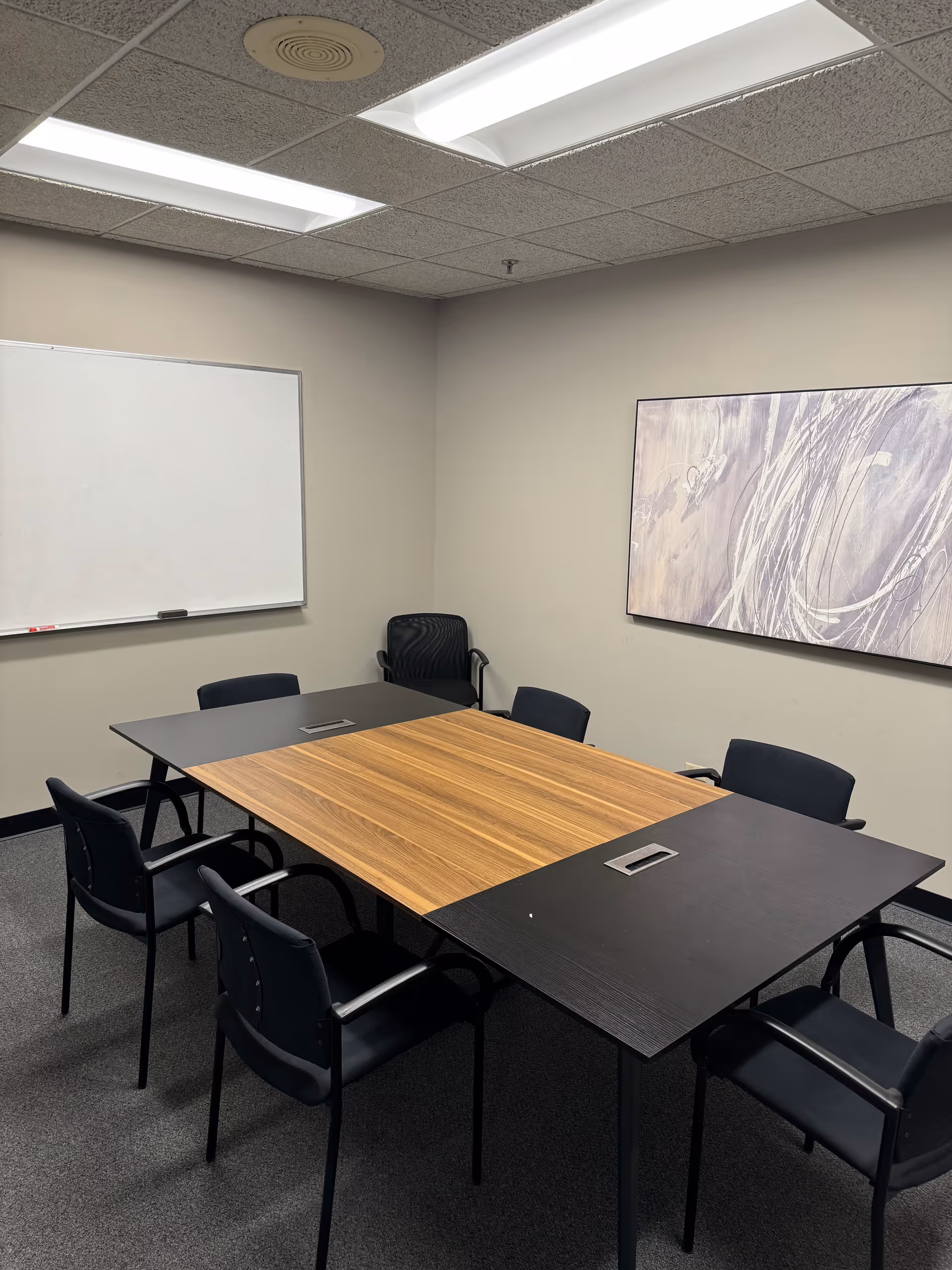 Conference room with a wooden center table, black chairs, whiteboard, and abstract wall art.