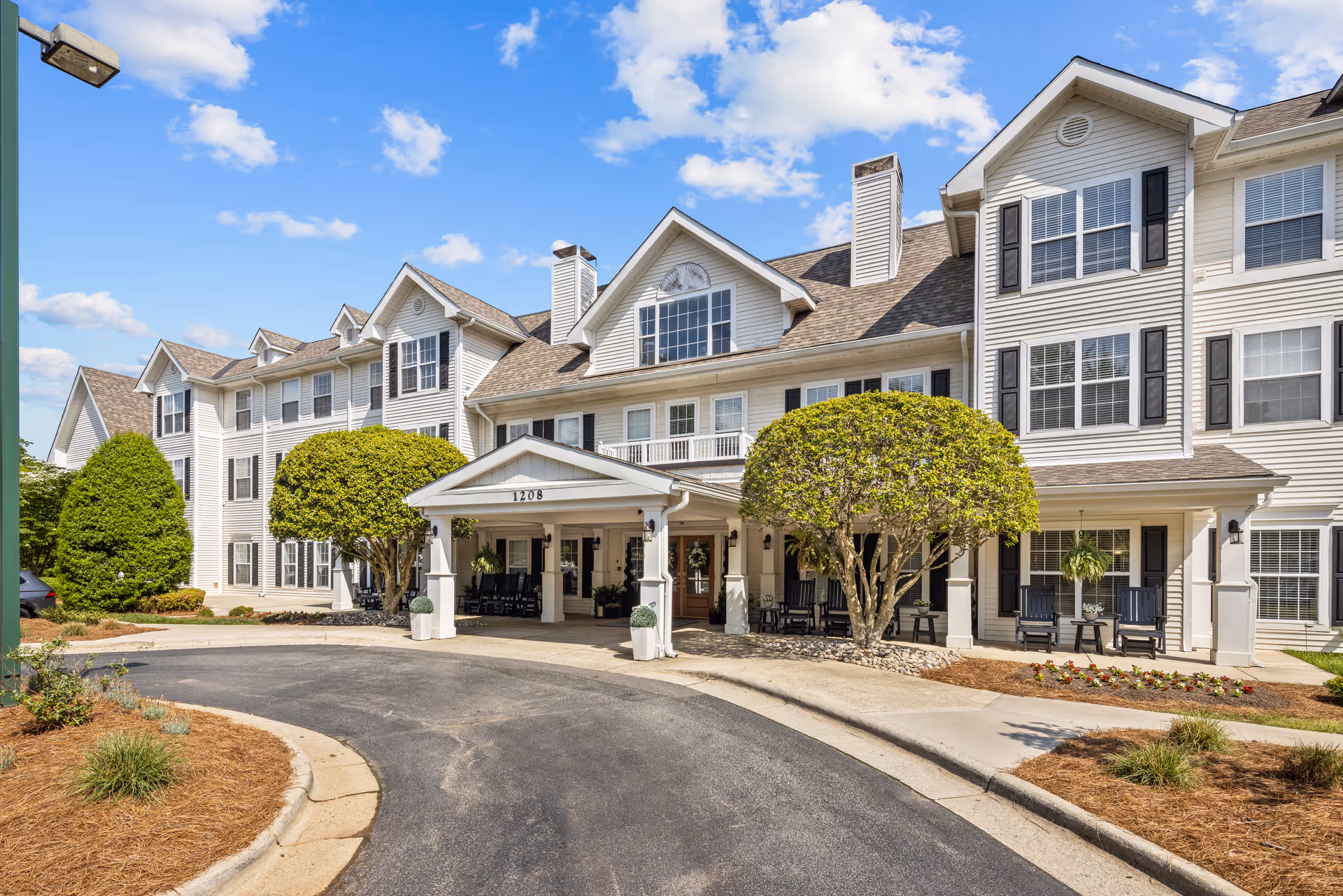 Exterior front view of TerraBella Greensboro senior living facility with a covered entrance, neatly trimmed trees, and a clear blue sky with scattered clouds.
