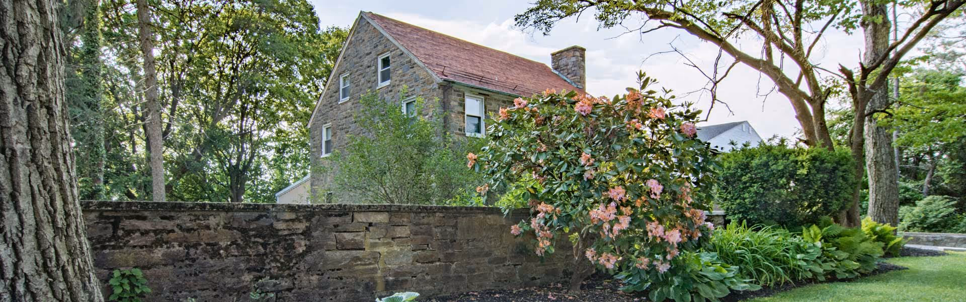Stone building with multiple windows partially obscured by a stone wall and lush greenery including a flowering bush and large trees under a partly cloudy sky.